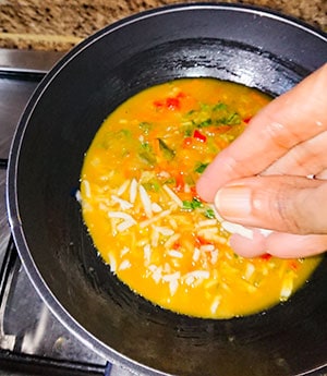 Grated cheese being sprinkled on cabbage omelette in a frying pan.