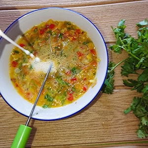 Beaten eggs with milk and vegetables in a bowl with a whisk in it.