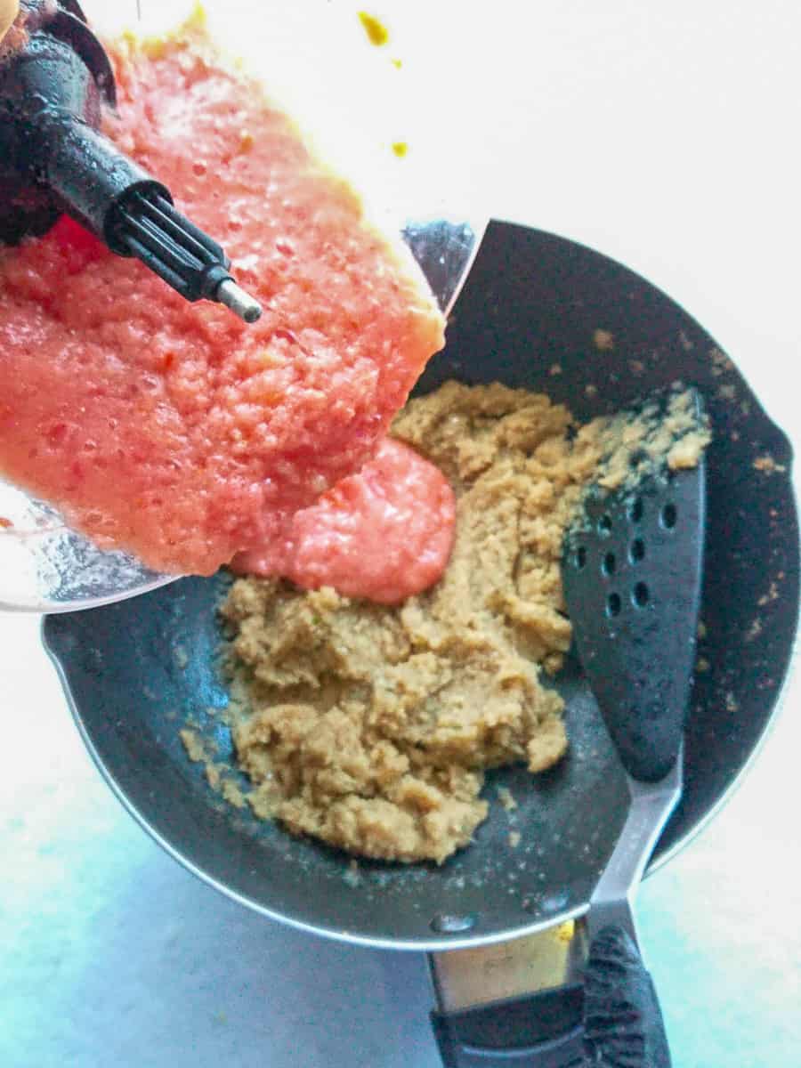 Tomato puree being added from a food processor jar into sauteed onion puree in a wok pan.