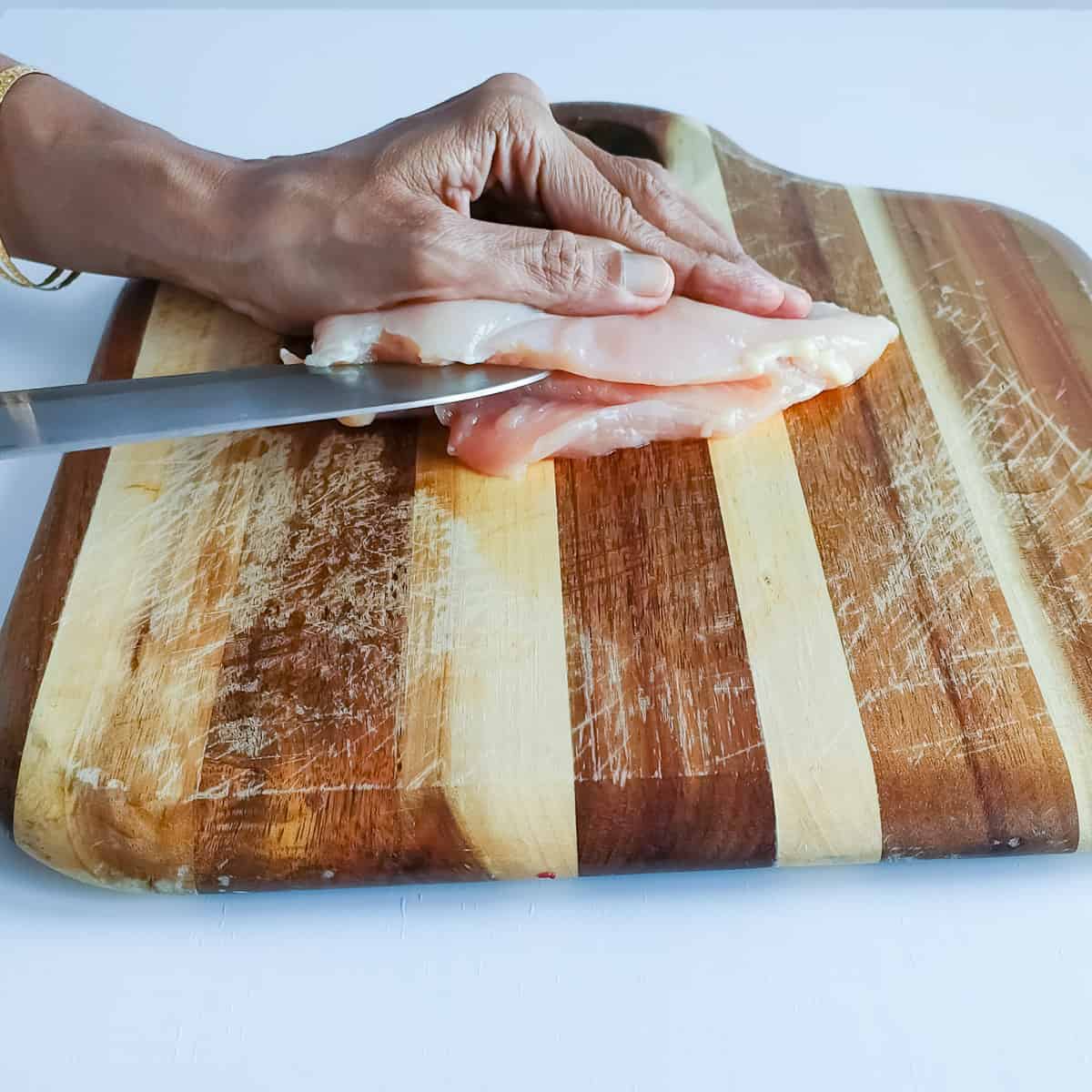 Chicken breast being thinly sliced with a knife on a chopping board.