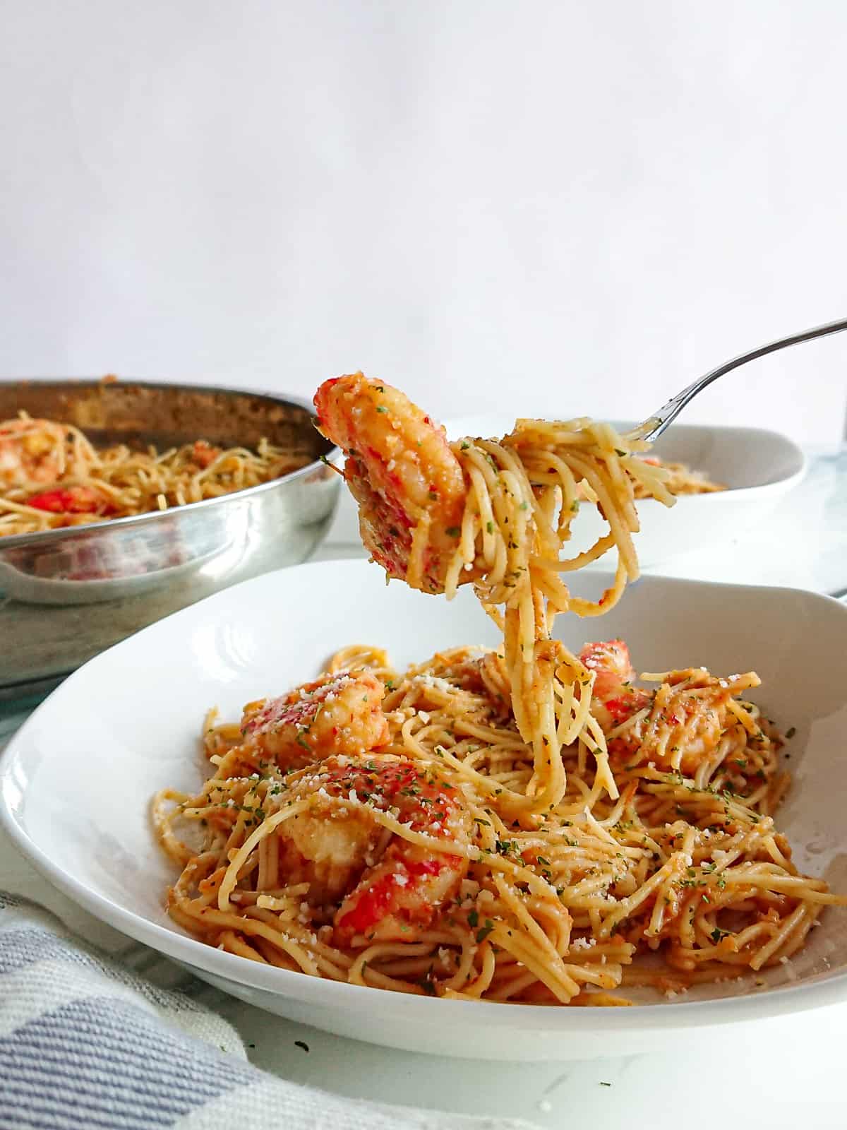 closeup view of tomato chilli prawn pasta on a white plate and a pan full of pasta in the background.