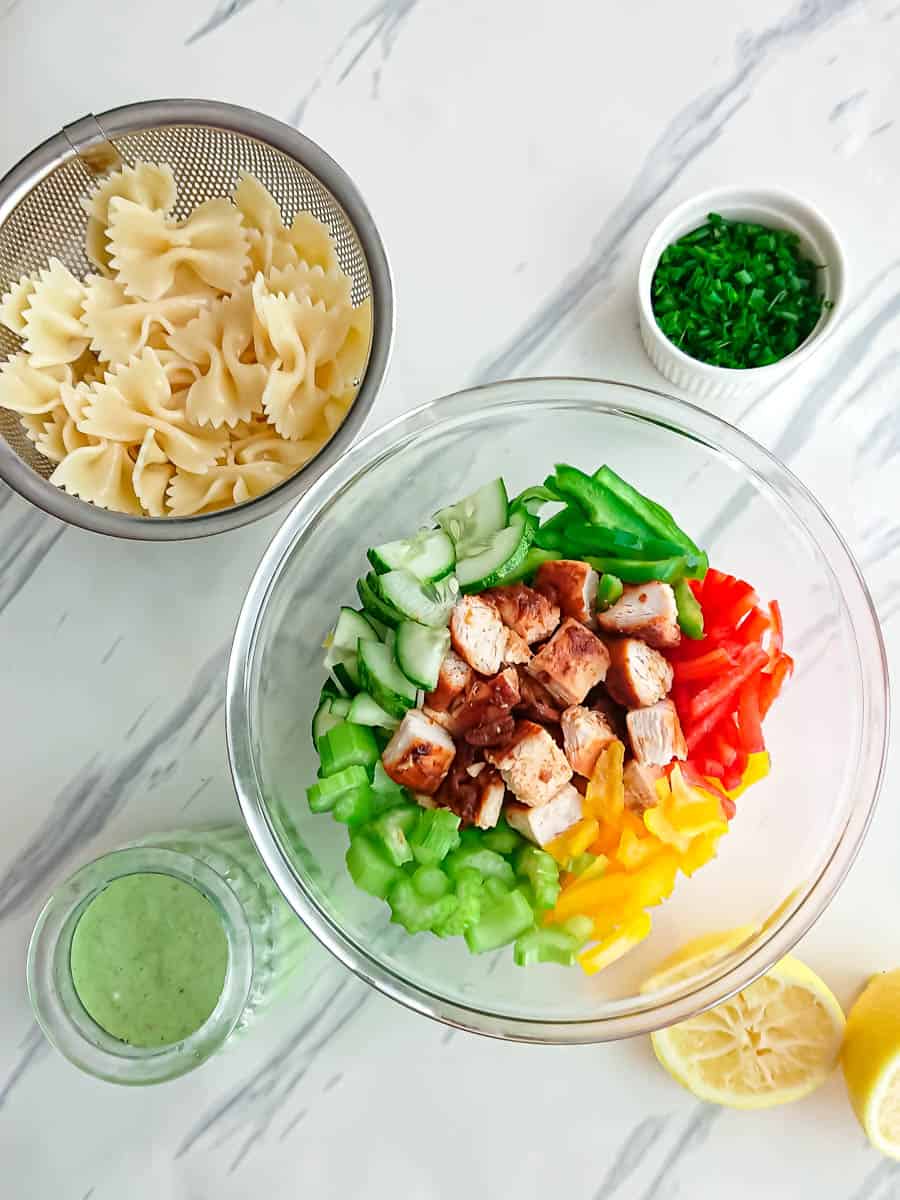 Ingredients for high protein pasta salad in a glass bowl, drained pasta in a strainer, lemon yogurt dressing in a jar and green onions in a bowl.