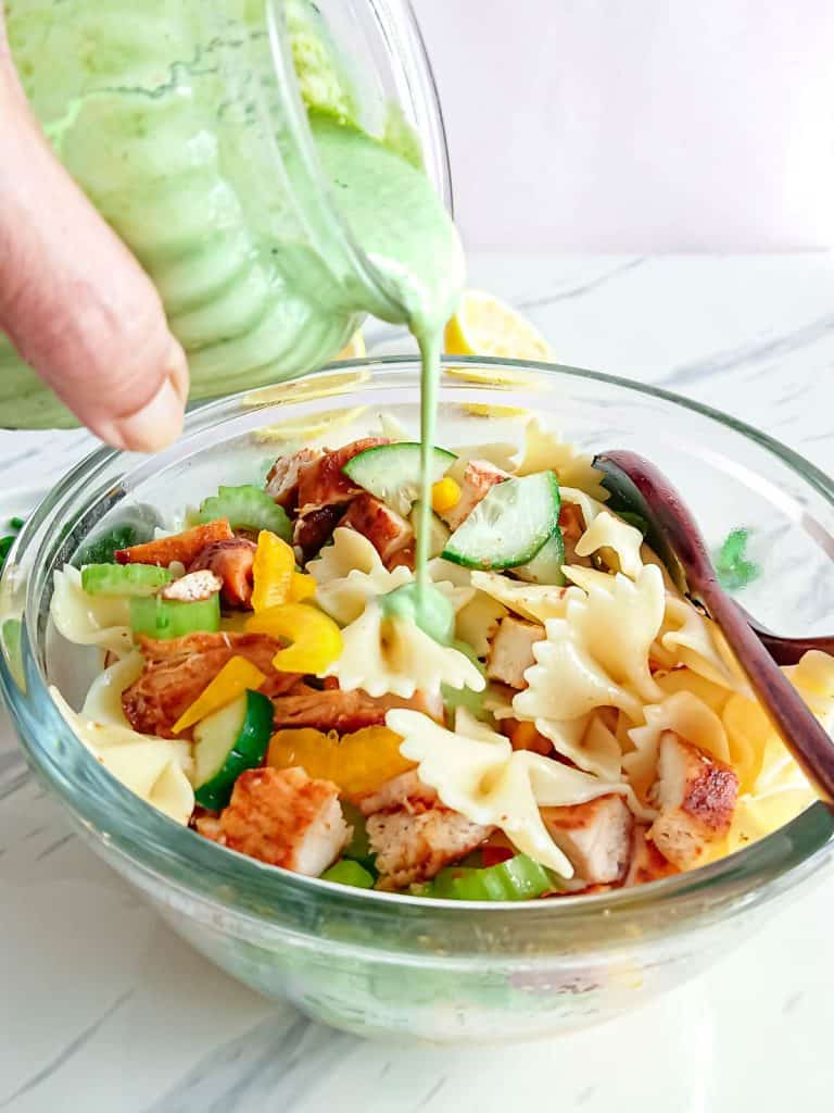 half of the creamy dressing being poured on chicken pasta salad in a glass bowl.