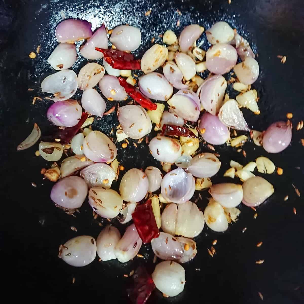 Shallots, chilies, cumin, garlic and ginger being sauteed in oil in a pan.