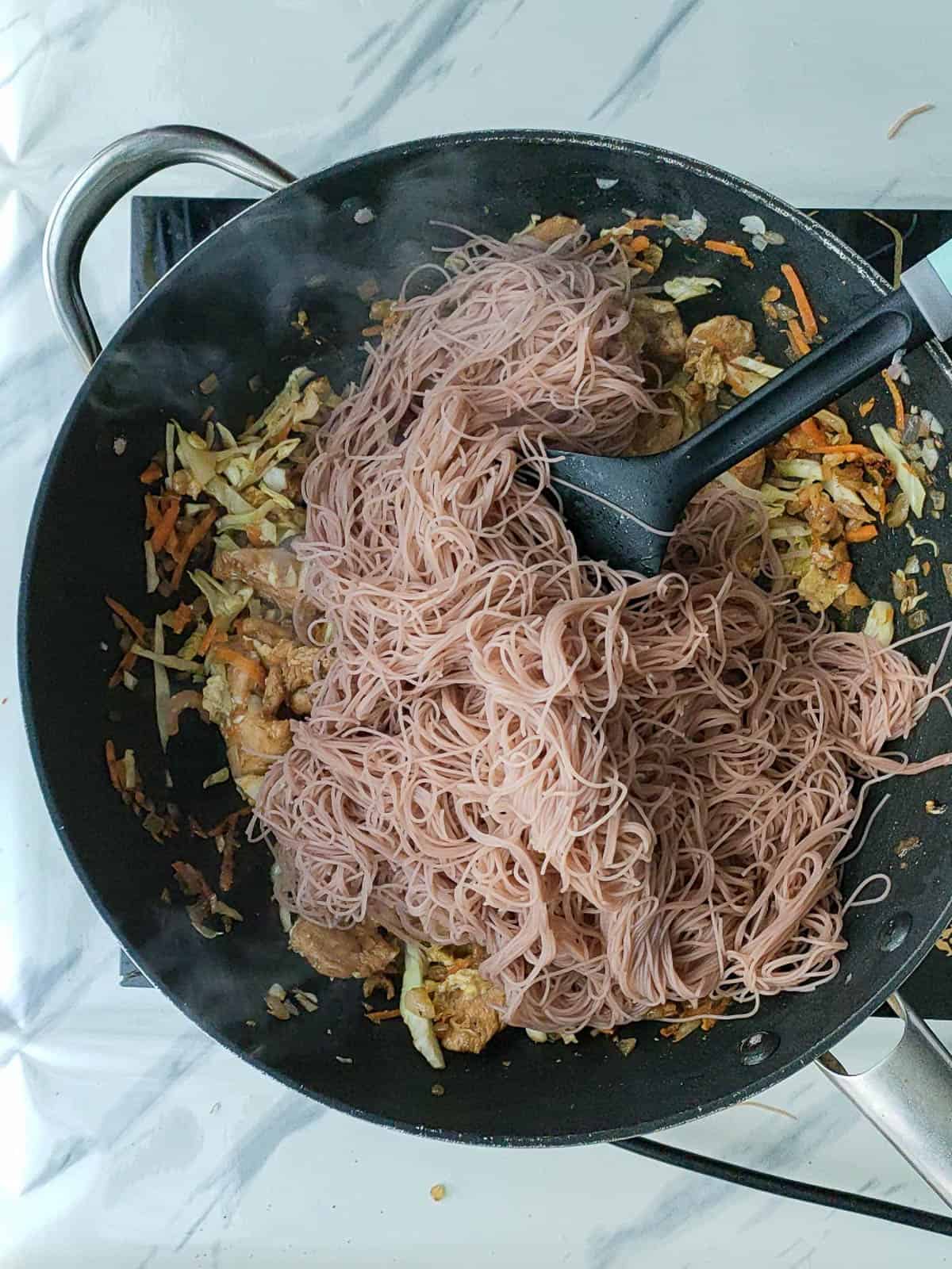 Rice vermicelli being stir-fried with vegetables in a non-stick wok.