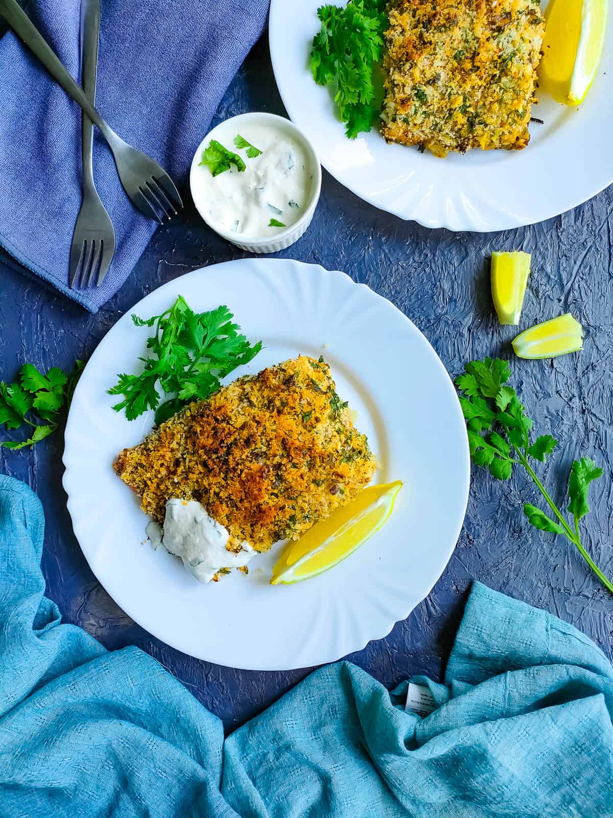 Panko baked cod served with yogurt sauce and garnished with parsley and lemon slices in 2 white plates.