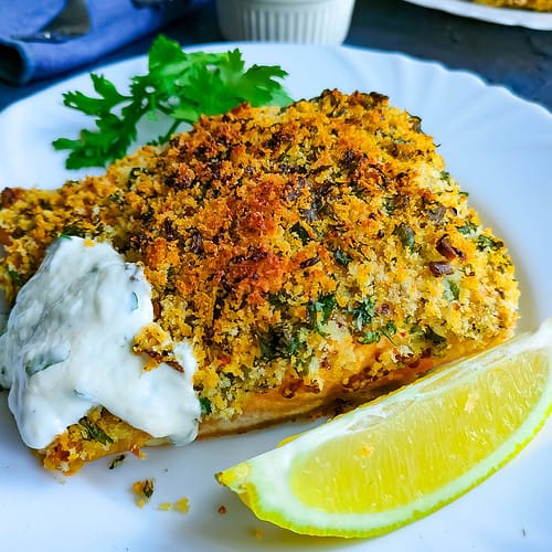 Baked Panko cod with yogurt sauce and garnished with parsley and lemon slices in a white plate.