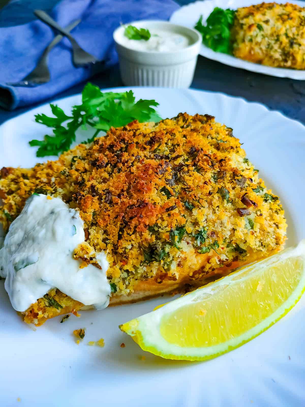 Baked panko fish with yogurt sauce and garnished with parsley and lemon slices on a white plate.