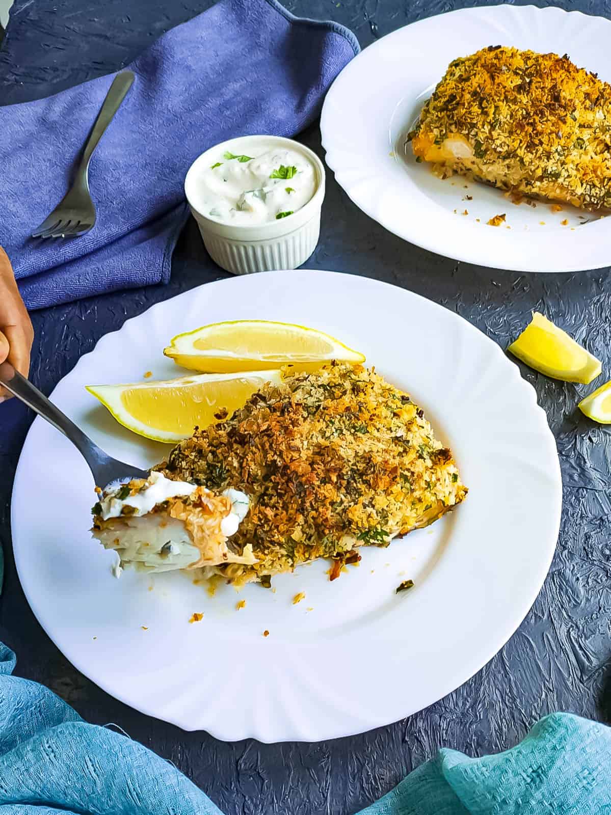 Baked panko crumbed fish on 2 white plates with yogurt sauce in a bowl next to them.