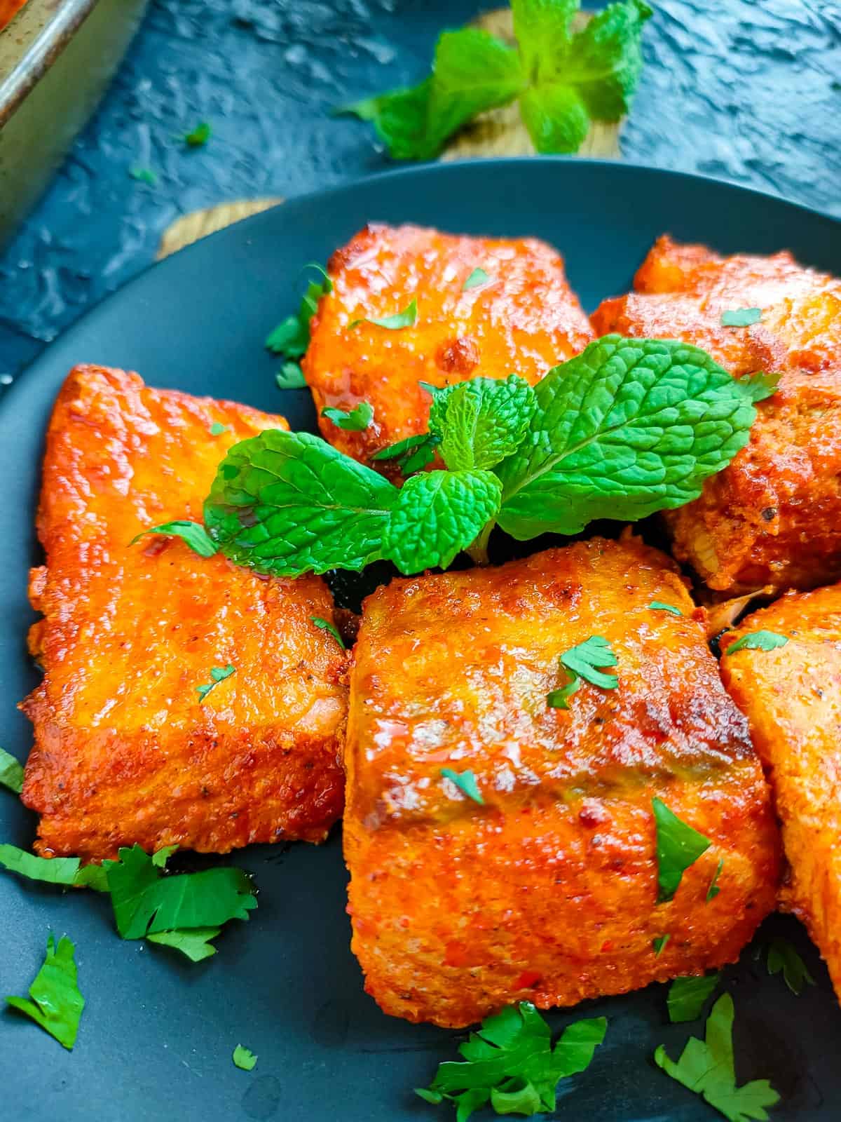 Closeup view of tandoori salmon tikka garnished with mint leaves on a black plate.