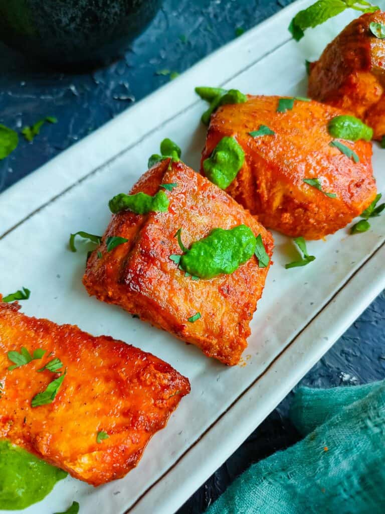 Tandoori salmon tikka on a white plate with mint sauce drizzled on them.