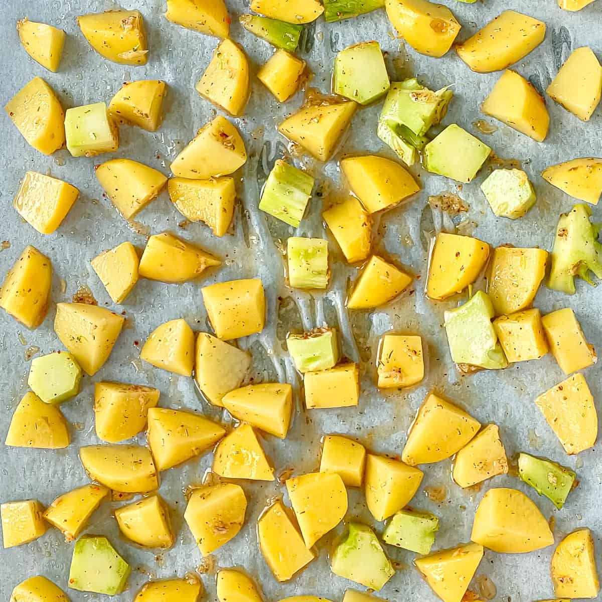 Cubed and seasoned potatoes and broccoli stems on a sheet pan lined with parchment paper.