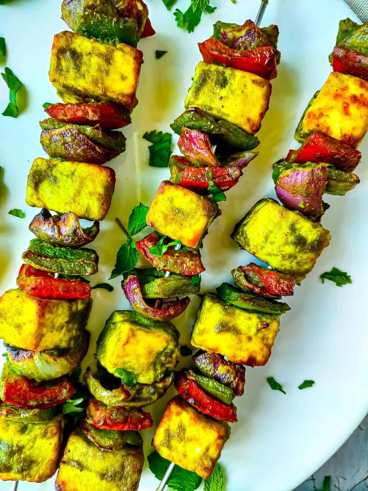 Hariyali Paneer Tikka (Oven roasted/ Pan fried) Hariyali paneer tikka on skewers served on a white plate.
