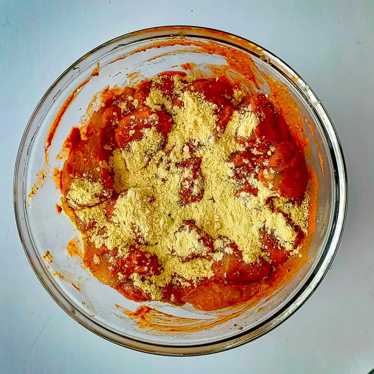 Roasted besan being added to marinated chicken cubes in a glass bowl.