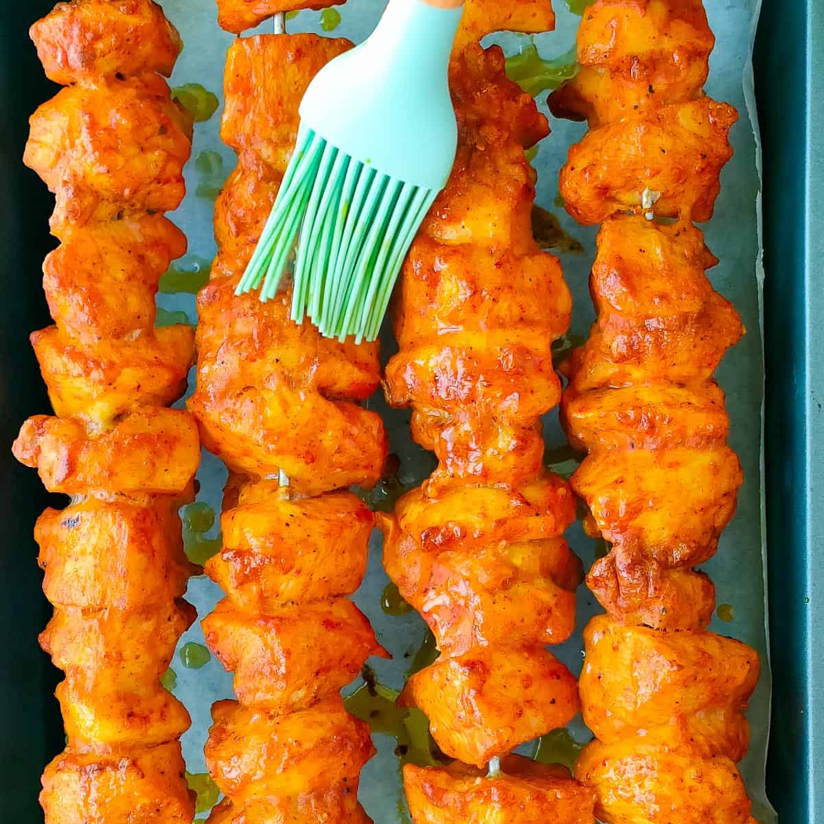 Tandoori chicken kebabs on a baking pan being basted with ghee using a brush.