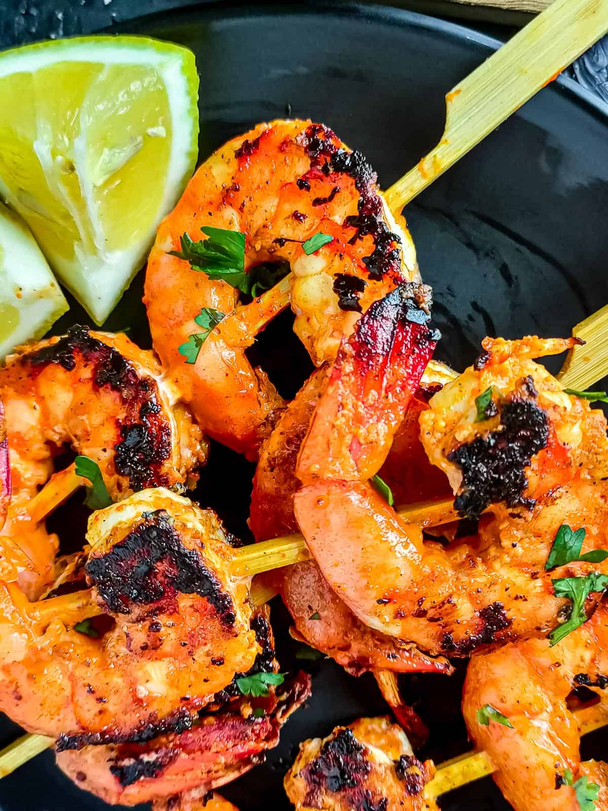 Tandoori king prawns on skewers on a black plate with lemon wedges.