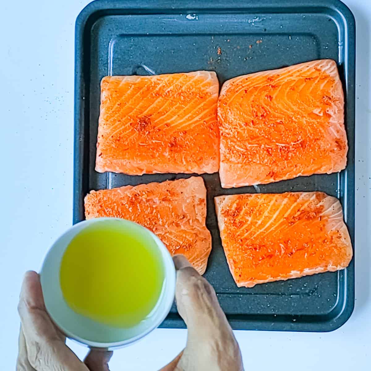 Lemon juice being drizzled on seasoned salmon fillets on a tray.