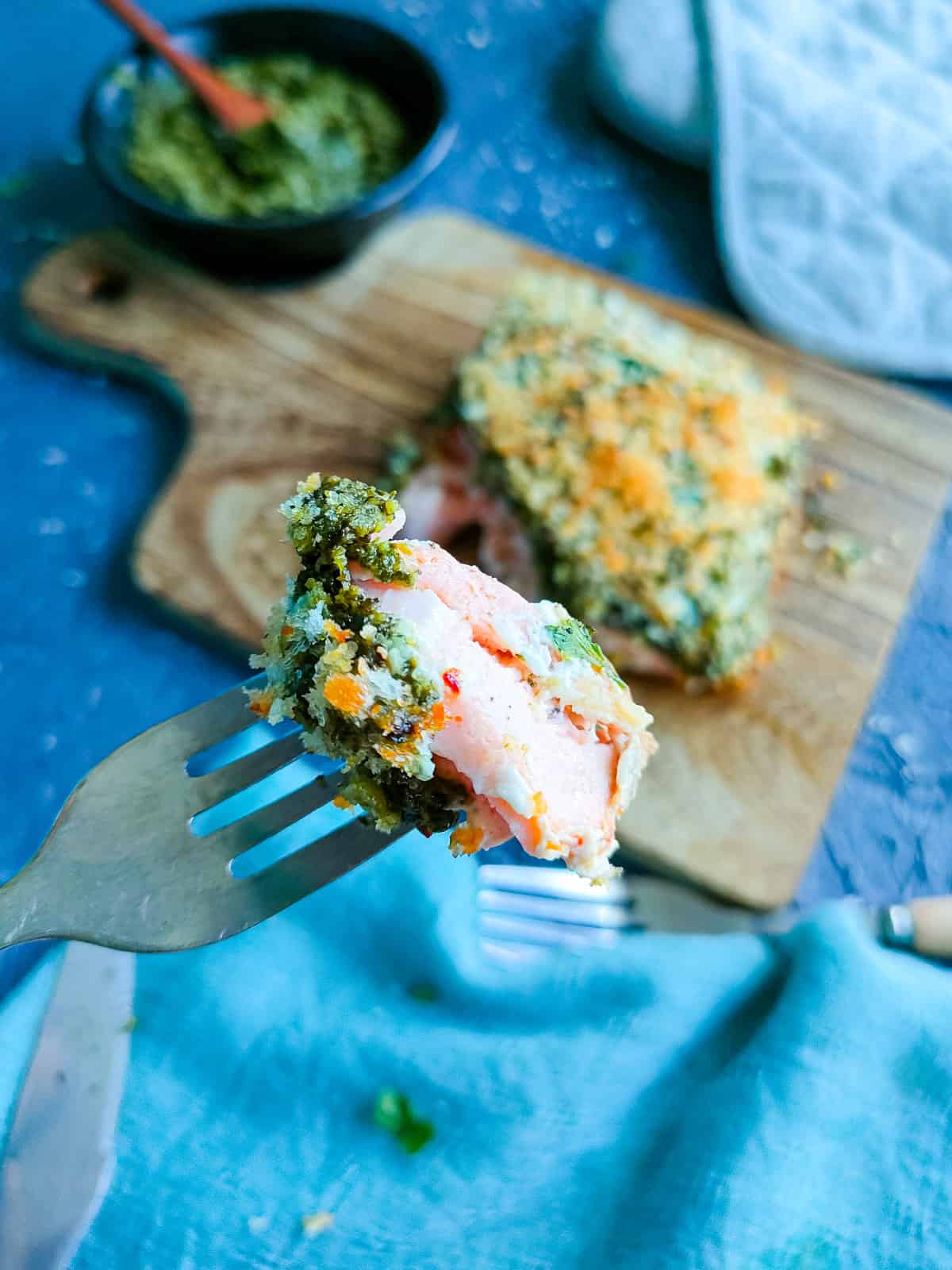 A piece of pesto crusted salmon on a fork with baked pesto salmon on a wooden board in the background.