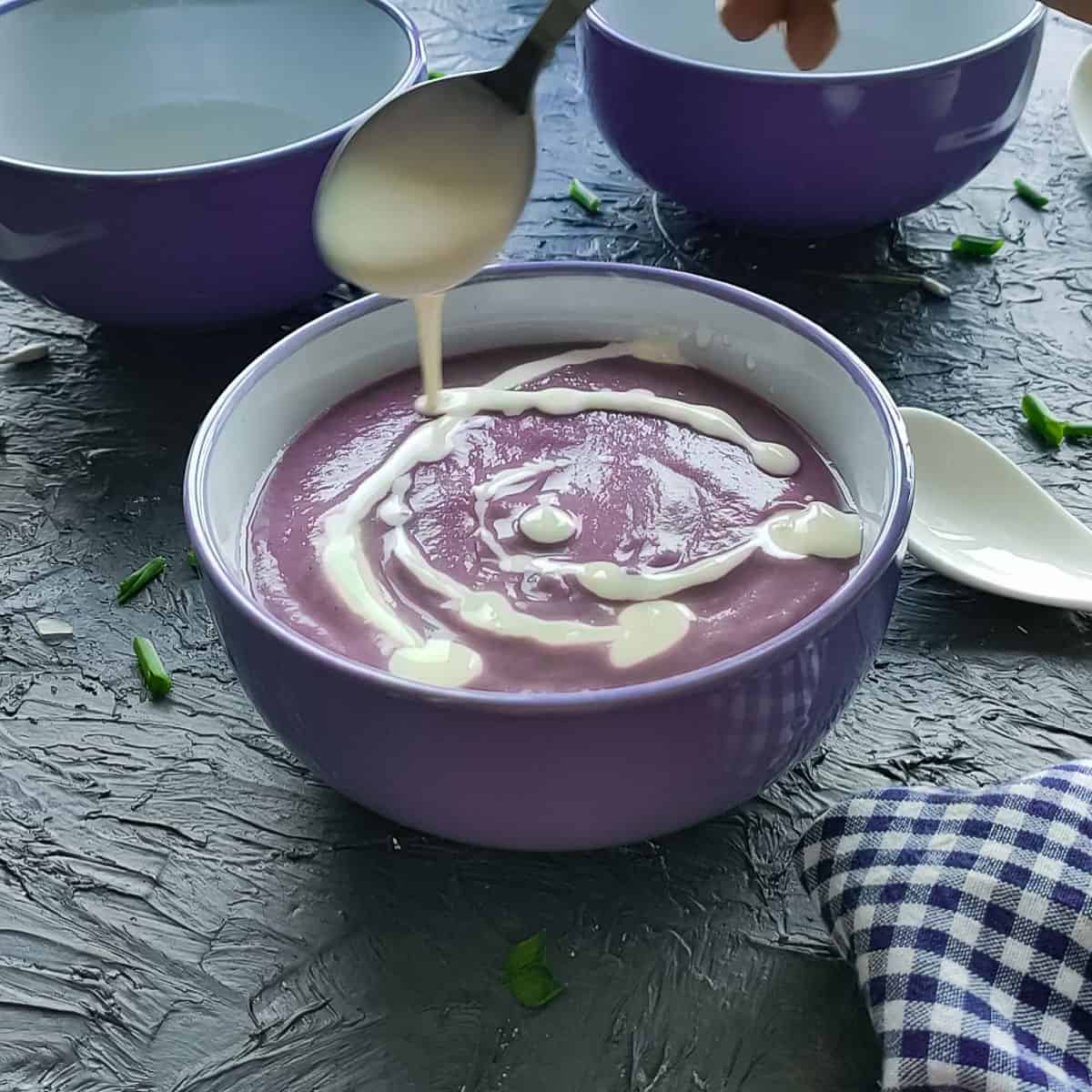 Yogurt with lemon juice being drizzled on red cabbage soup in a white bowl.