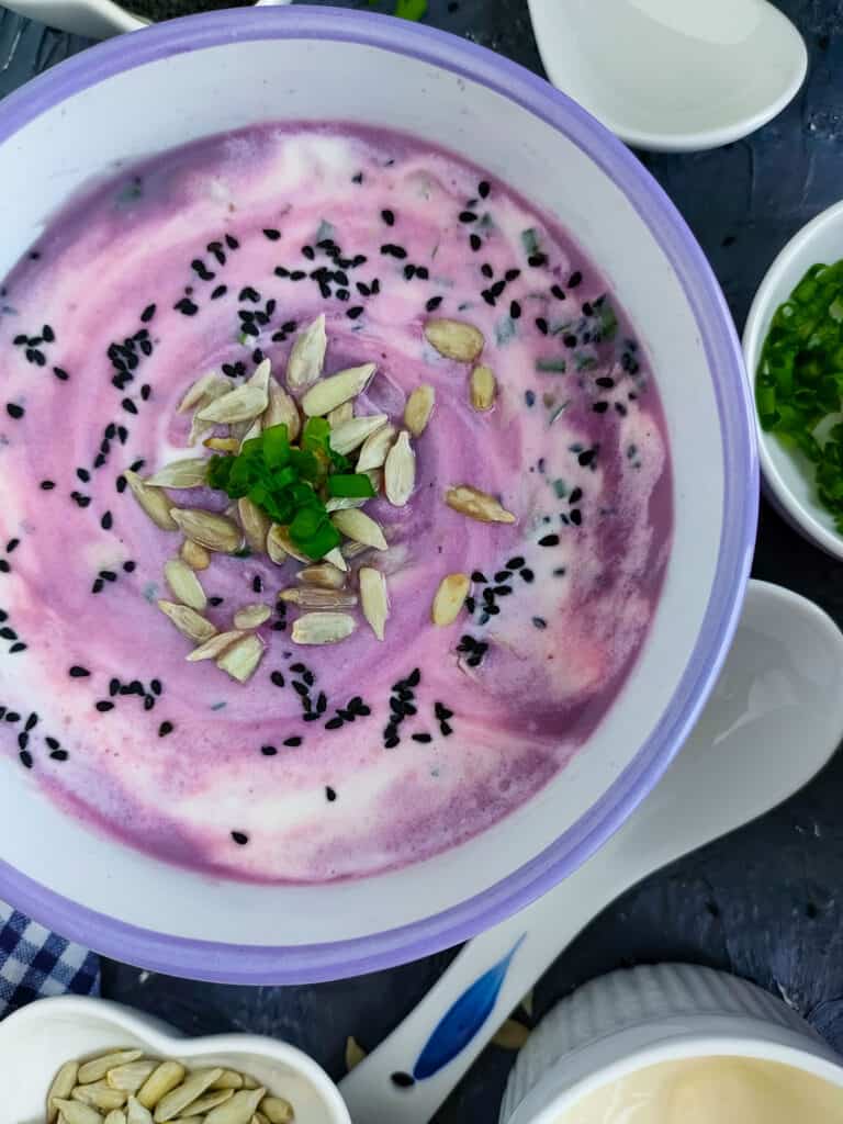 Red cabbage soup in a white and blue bowl with bowls of sunflower and nigella seeds, green onions, and yogurt next to it.