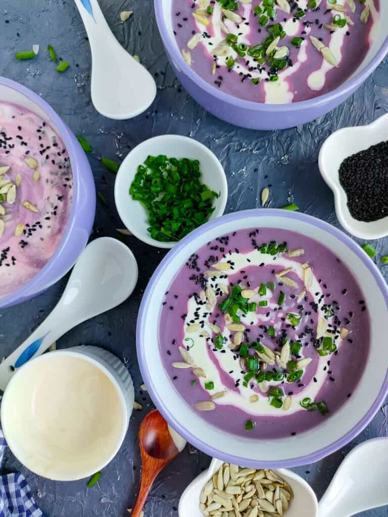 Red cabbage soup in 3 white and blue bowls with bowls of sunflower and nigella seeds, green onions, and yogurt next to them.