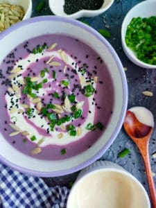 Red cabbage soup in a white and blue bowl with bowls of sunflower and nigella seeds, green onions, and yogurt next to it.