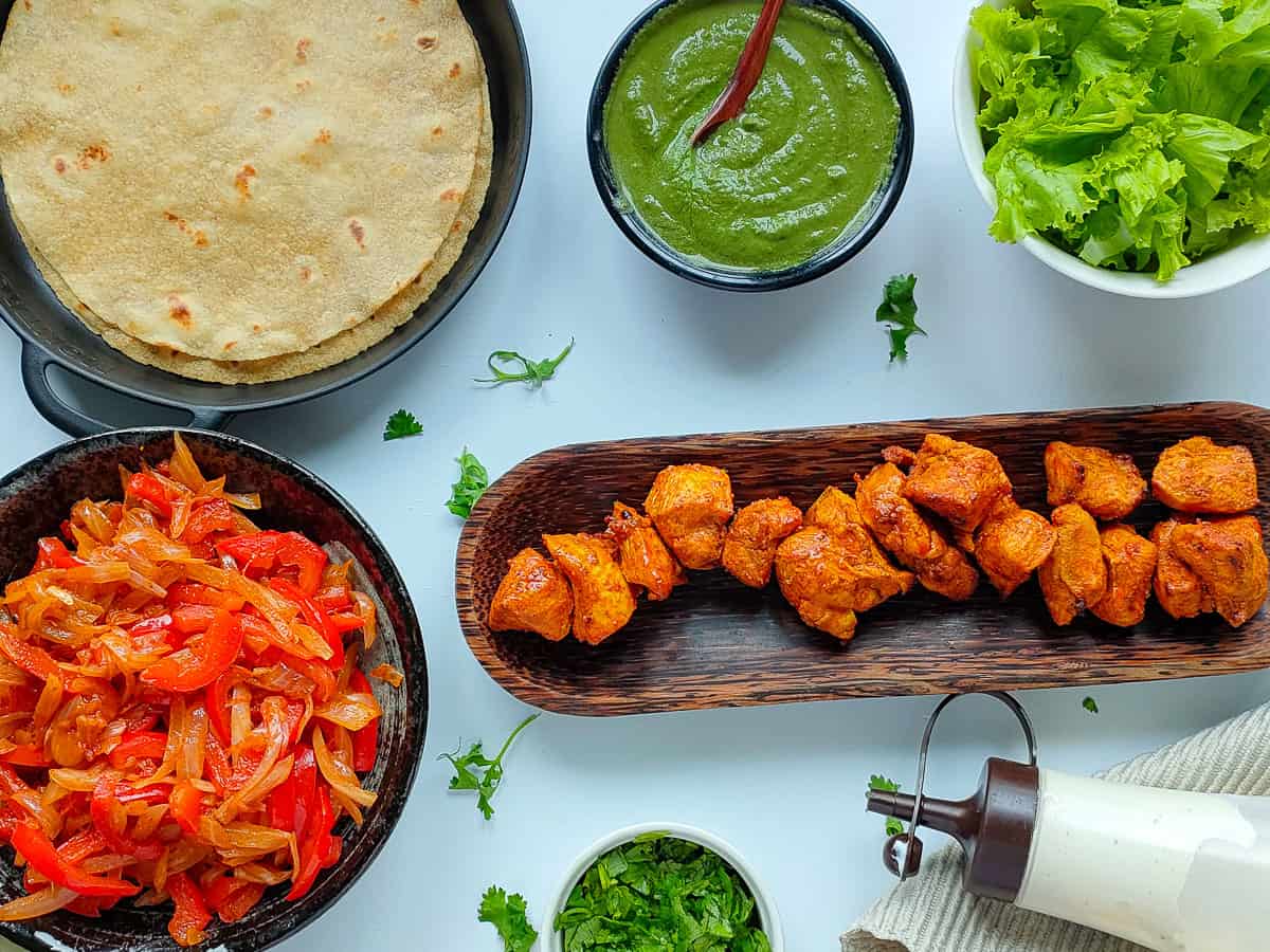 Tandoori chicken tikka, sauteed veggies, rotis, green chutney, lettuce, yogurt sauce, and coriander leaves for chicken tikka wraps in separate plates and bowls.