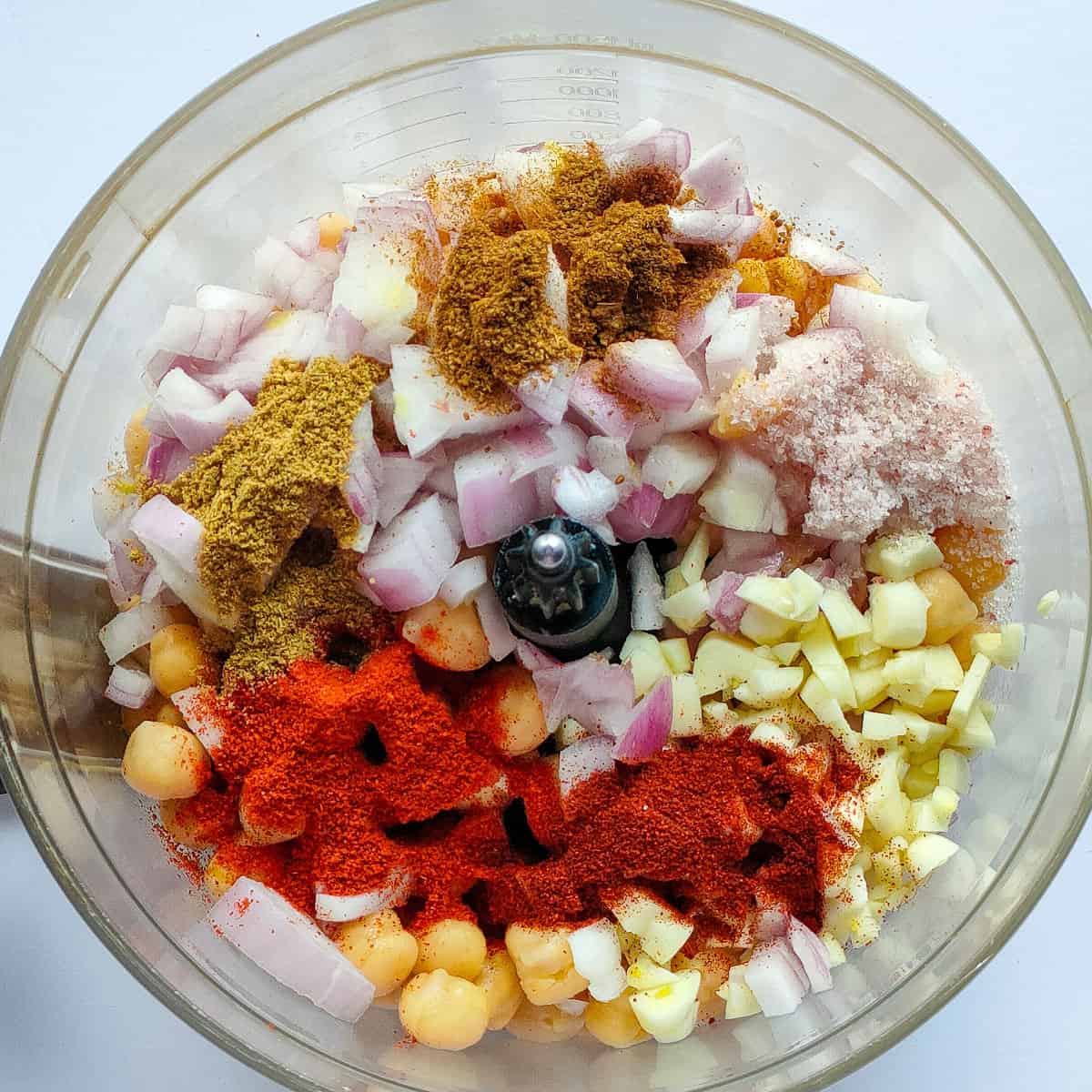 Chickpeas, onions, garlic and spices in a food processor jar ready to be processed.