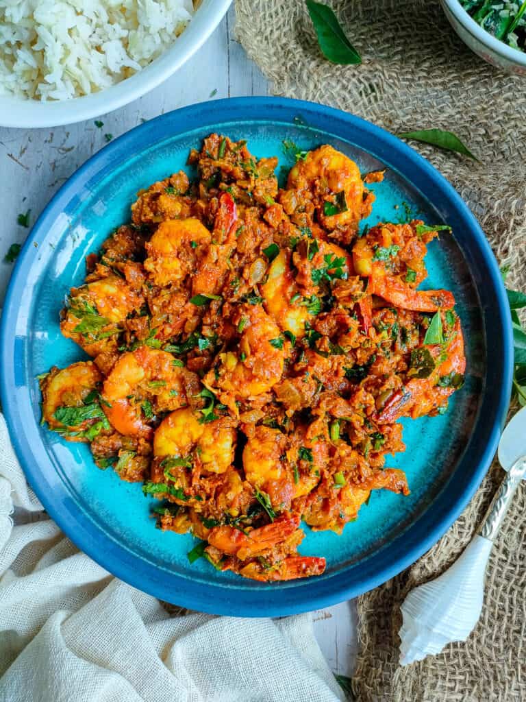 Prawn fry masala on a blue plate with a bowl of rice next to it.