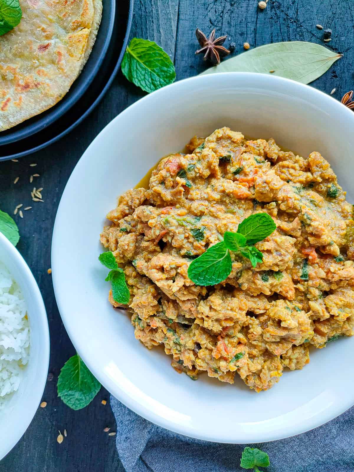 Minced chicken curry in a white bowl with a bowl of rice and rotis on a black plate next to it.