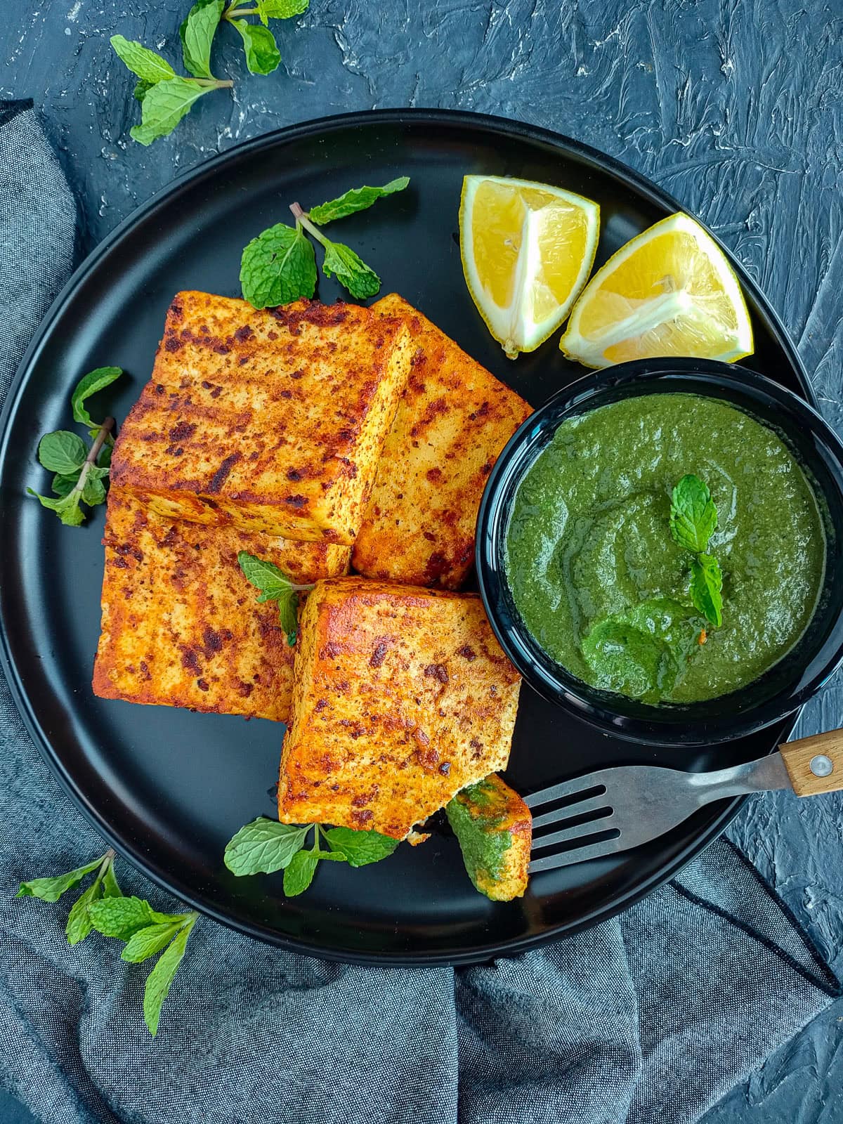 Tandoori paneer tikka with mint chutney and garnished with mint leaves and lemon wedges on a black plate.