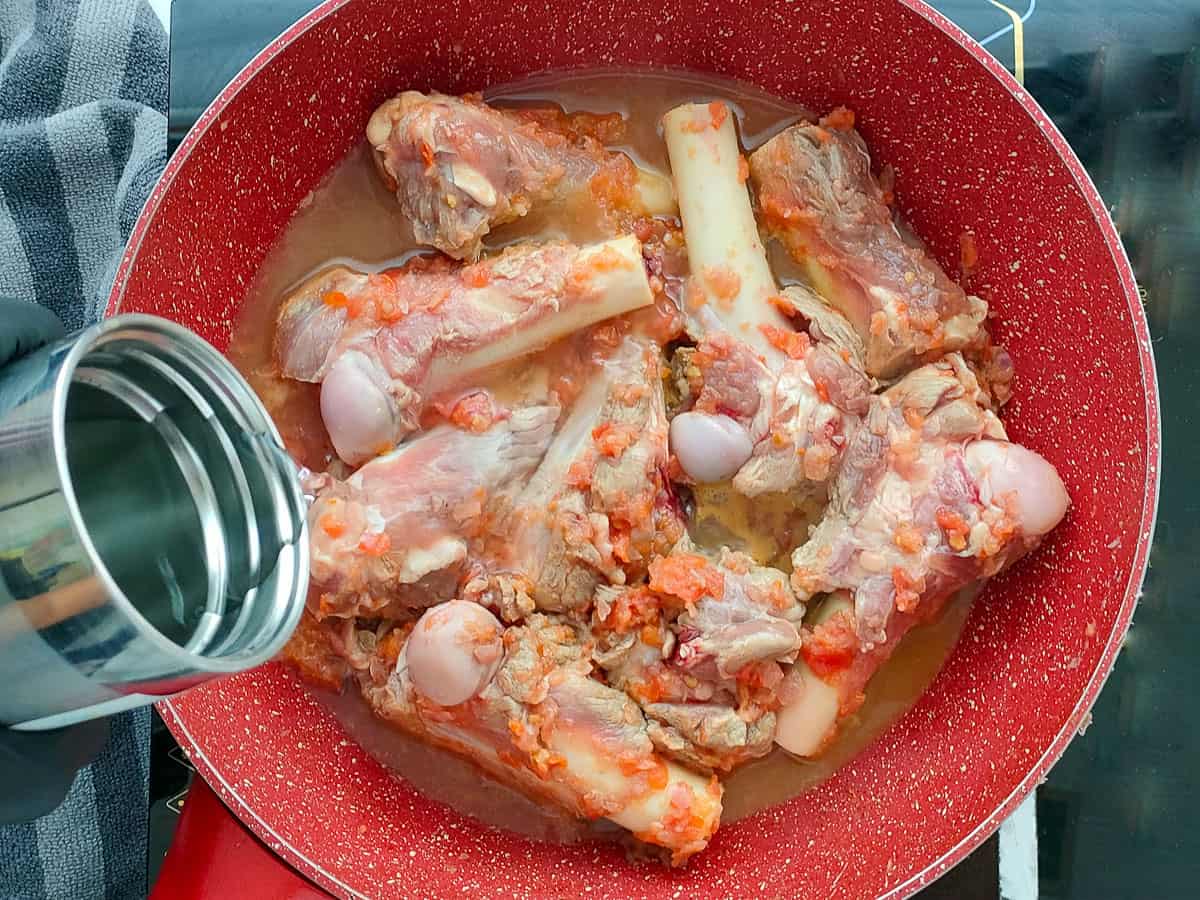 Water being added to mutton bones with onion tomato mixture in a pink wok pan.