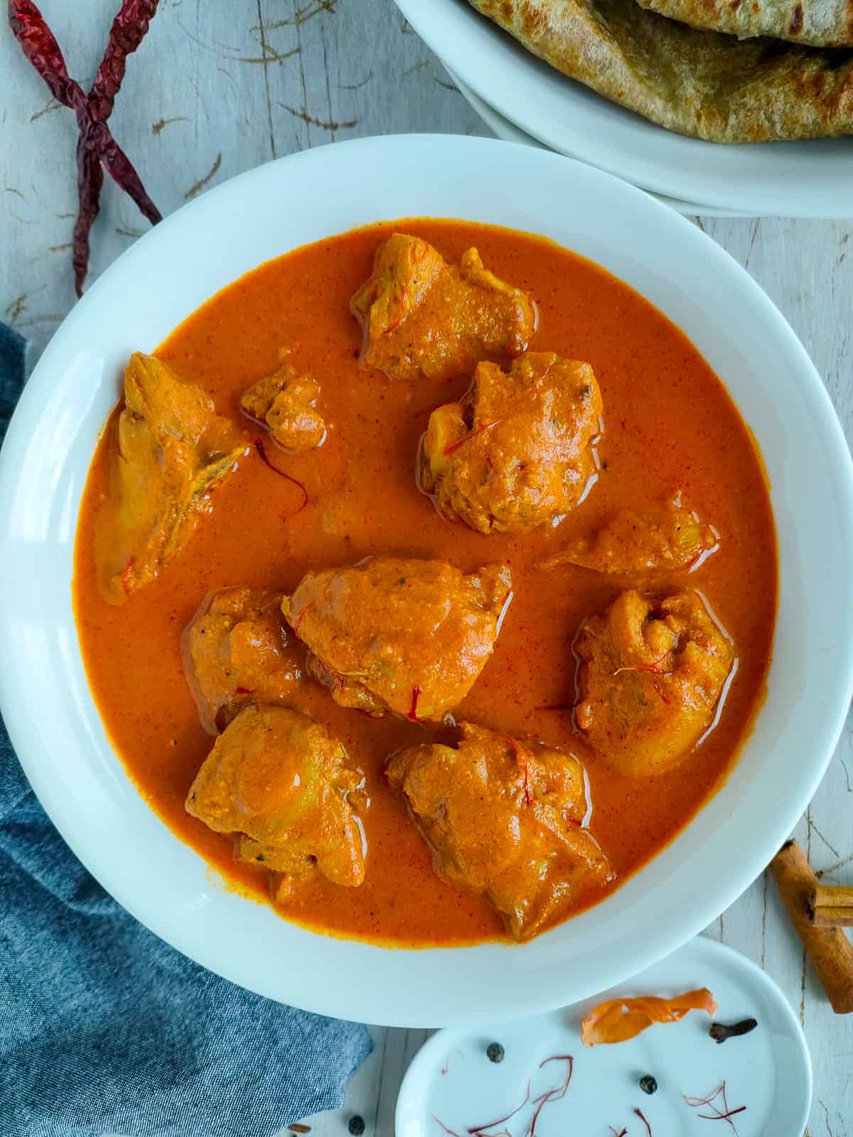Easy Chicken Rogan Josh Rogan josh chicken in a large white bowl with a plate of rotis next to it.