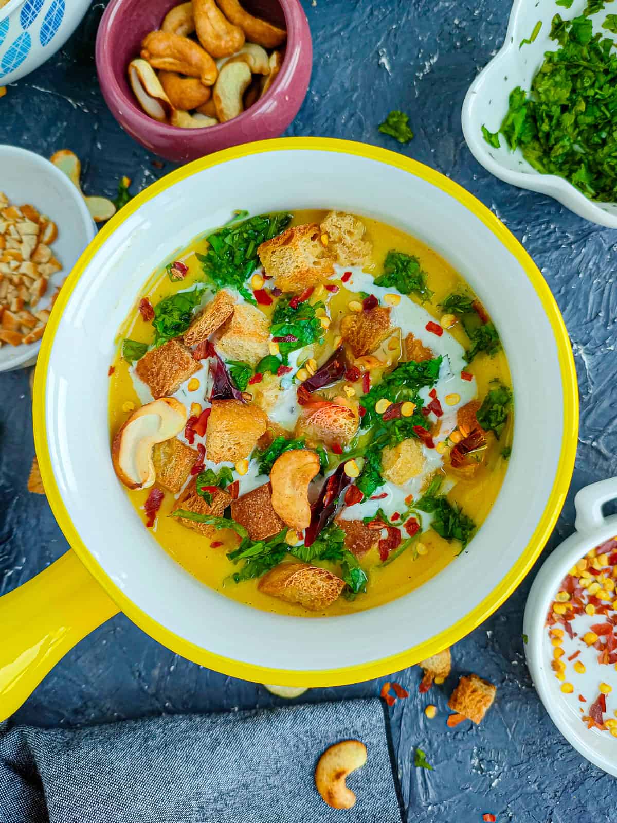 Spicy cauliflower low carb soup without cream in a white and yellow bowl and roasted cashews, chili flakes and coriander leaves in different bowls placed next to it.