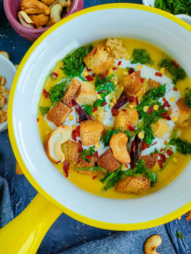 Spicy cauliflower cashew soup without cream in a white and yellow bowl and roasted cashews, chili flakes and coriander leaves in different bowls placed next to it.