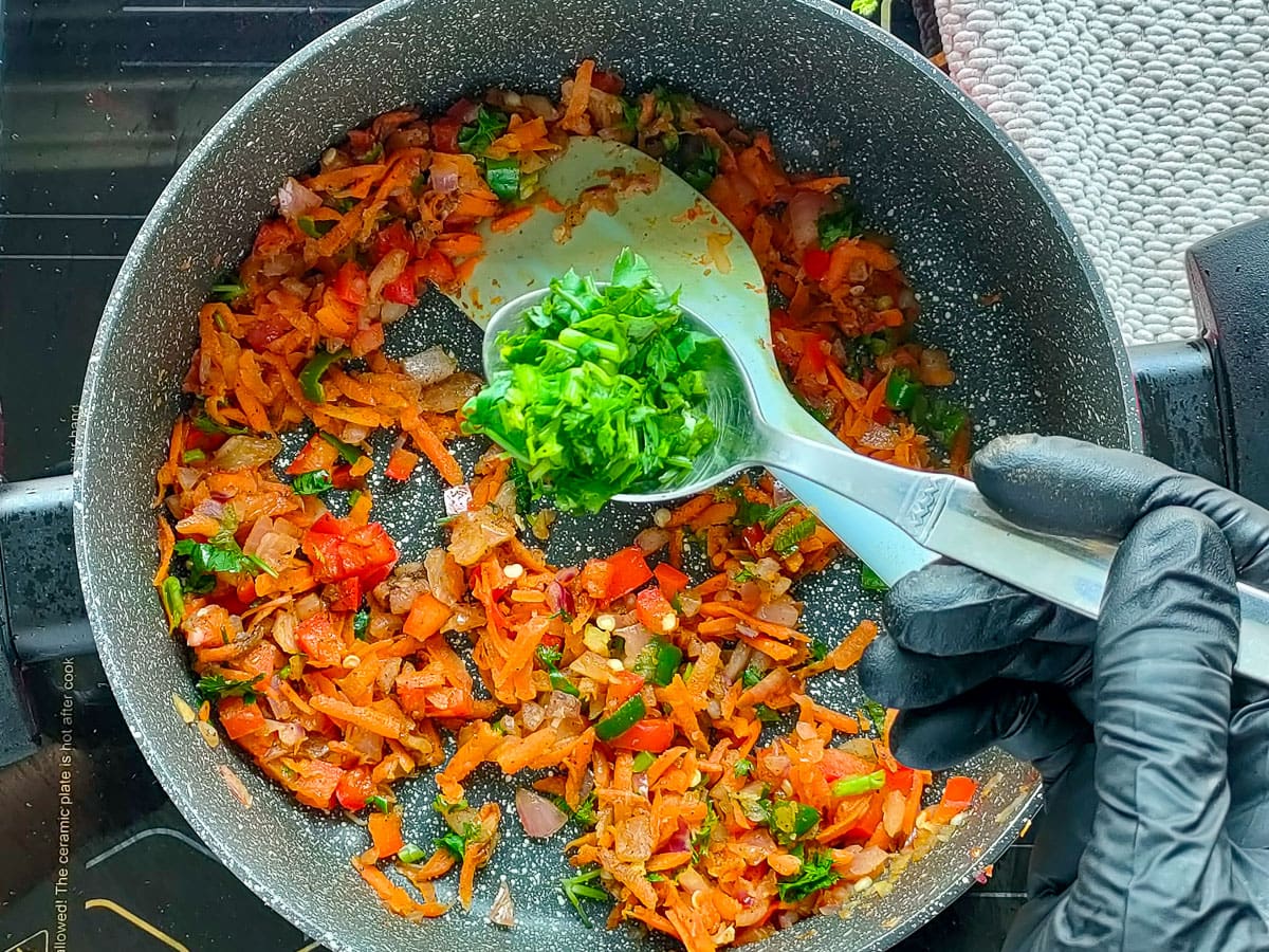 A spoon of chopped coriander leaves being added to sauteed vegetables in a pot.