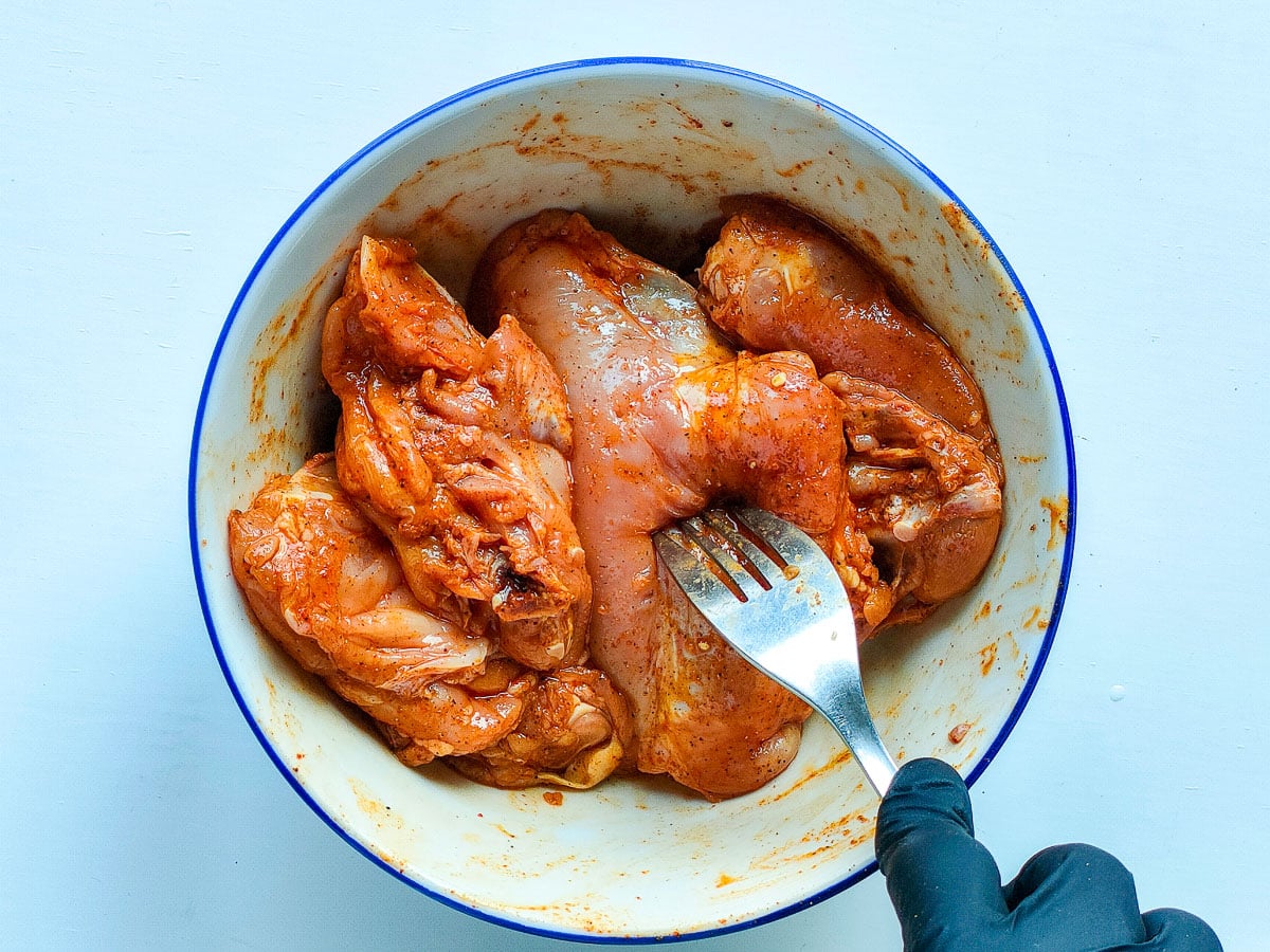 Marinated chicken thighs in a medium white bowl being pierced with a fork.