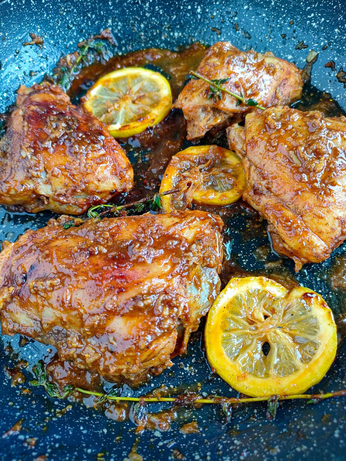 Chicken thighs with honey lemon pepper sauce garnished with thyme and caramelized lemon slices in a frying pan.