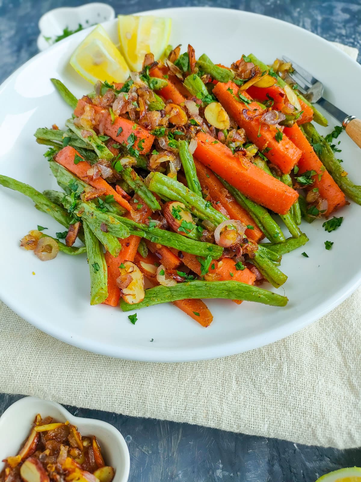Roasted green beans and carrots with toasted almond-shallot topping and lemon wedges on a white plate.