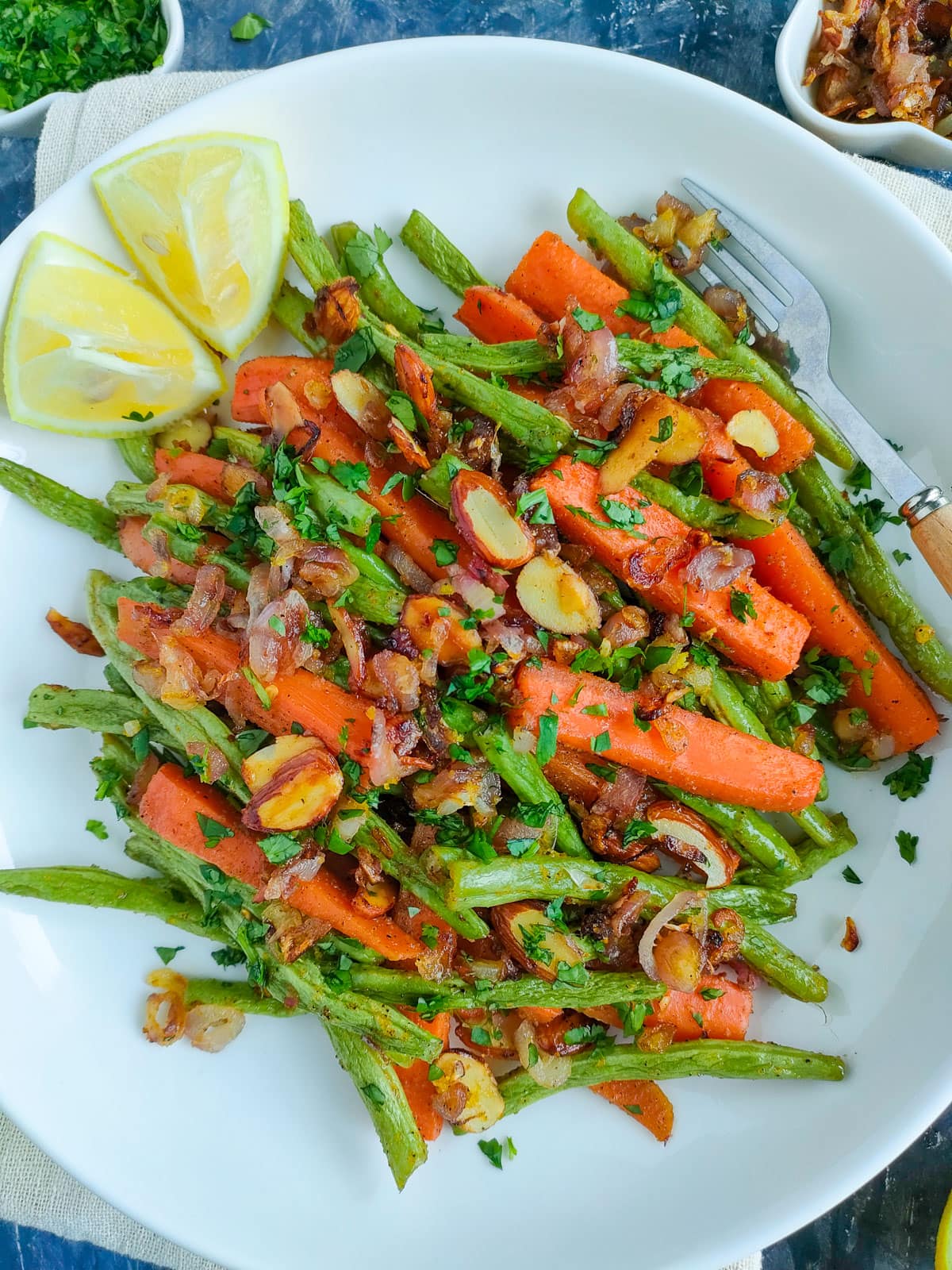 Roasted green beans and carrots with toasted almond topping and lemon wedges on a white plate.
