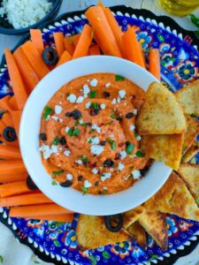 Feta paprika dip in a white bowl with carrot sticks and pita chips on a plate.