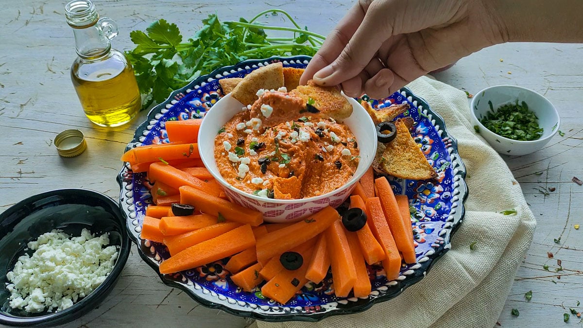 Feta paprika dip being scooped out with pita chips from a white bowl.
