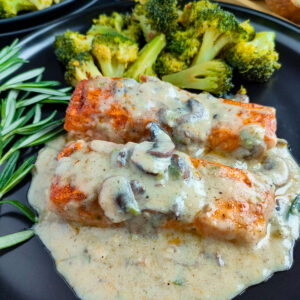 Salmon with mushroom cream sauce on a black plate with steamed broccoli and garnished with rosemary.