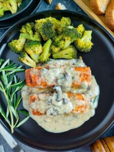 Salmon in creamy mushroom sauce on a black plate with steamed broccoli and garnished with rosemary.