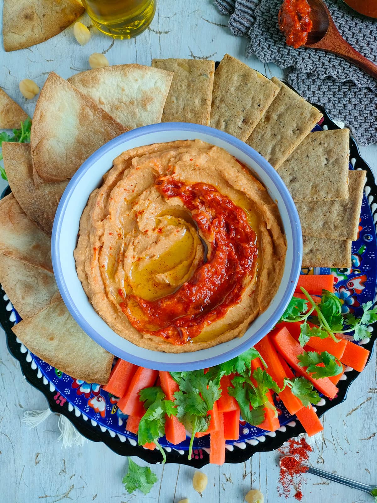 Harissa hummus in a bowl with fresh vegetables, pita chips and crackers on a platter.