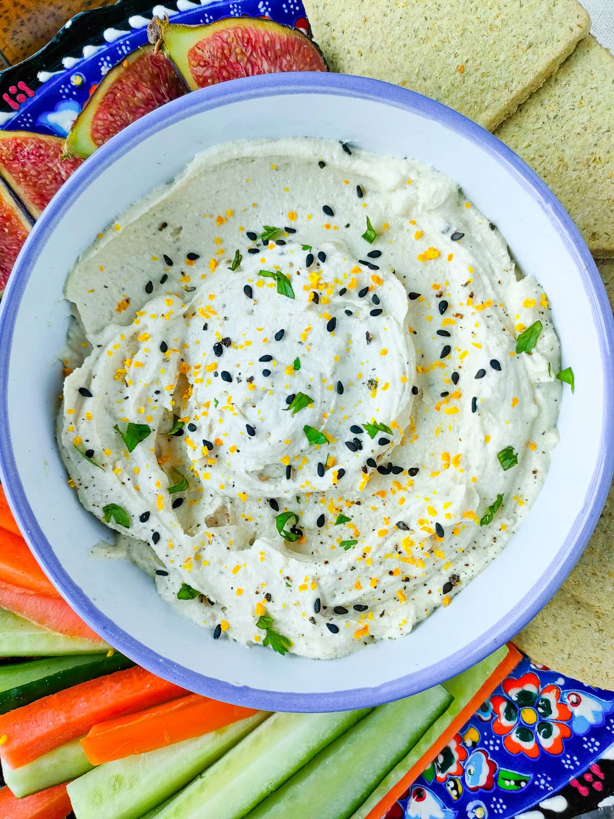 Greek yogurt tahini sauce in a blue and white bowl with vegetable sticks, fruits and crackers on a plate.