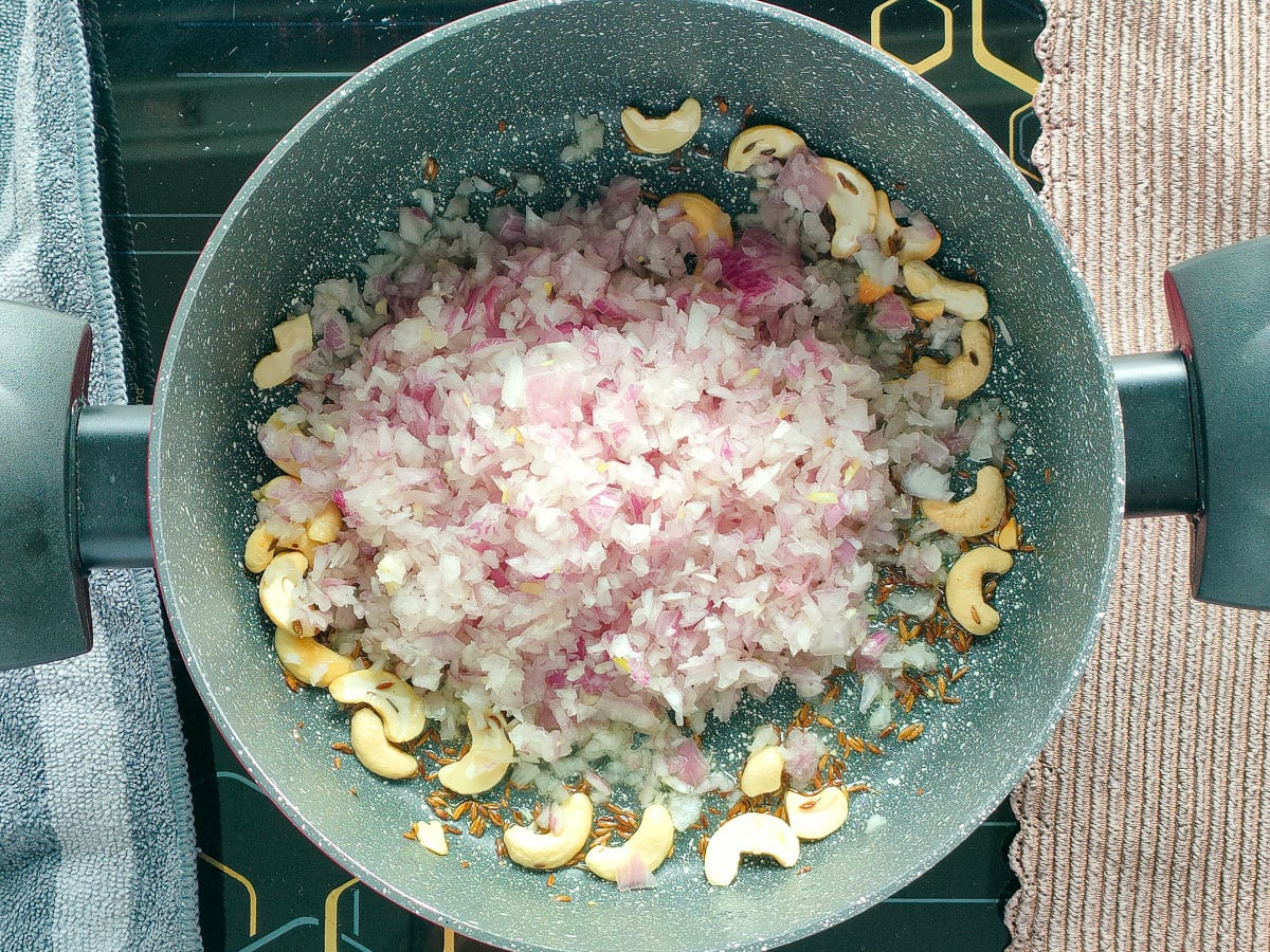 Onions with Cumin seeds and cashew nuts being fried in a pot.