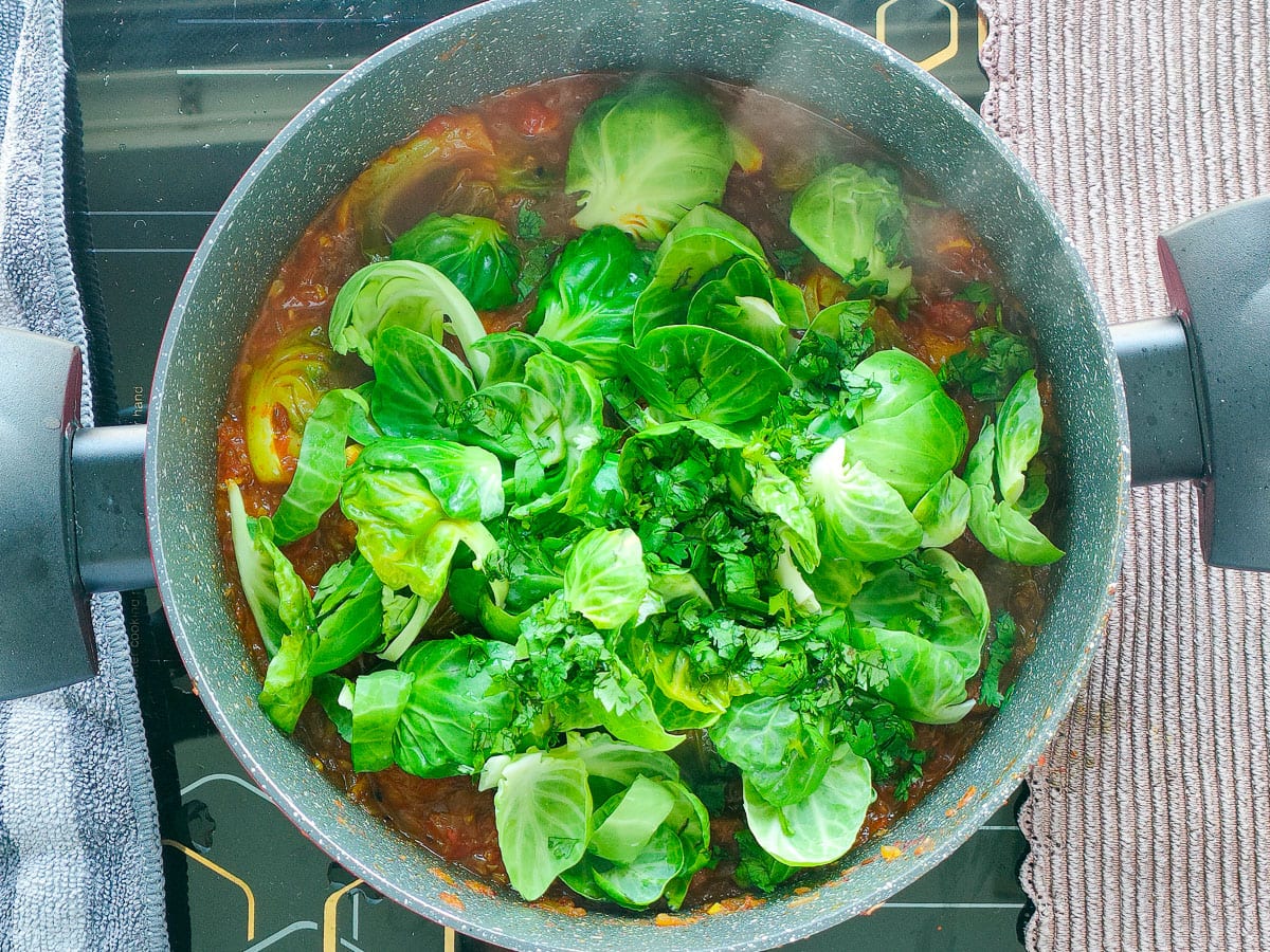 Loose leaves of Brussels sprouts and coriander leaves being added to the simmering curry in a pot.