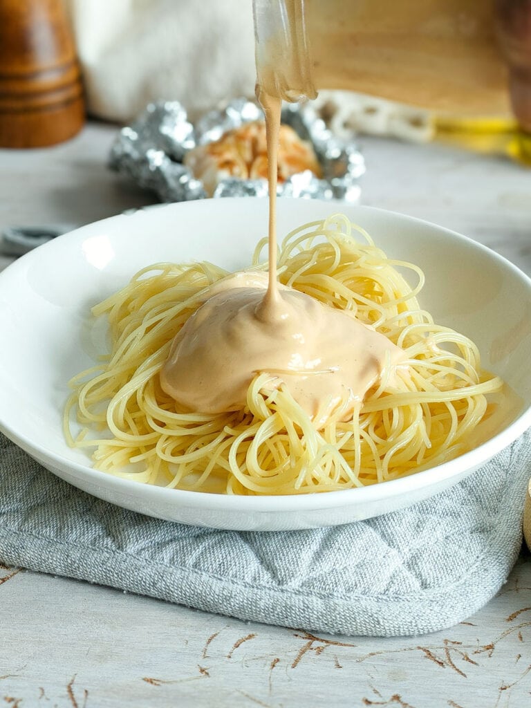 Roasted garlic pasta sauce being drizzled on pasta in a white plate.