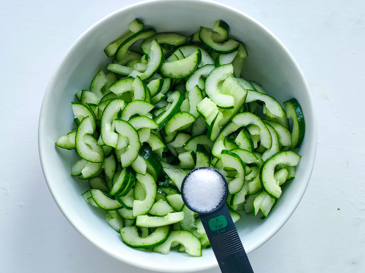 Salt being added to sliced cucumbers in a large bowl.